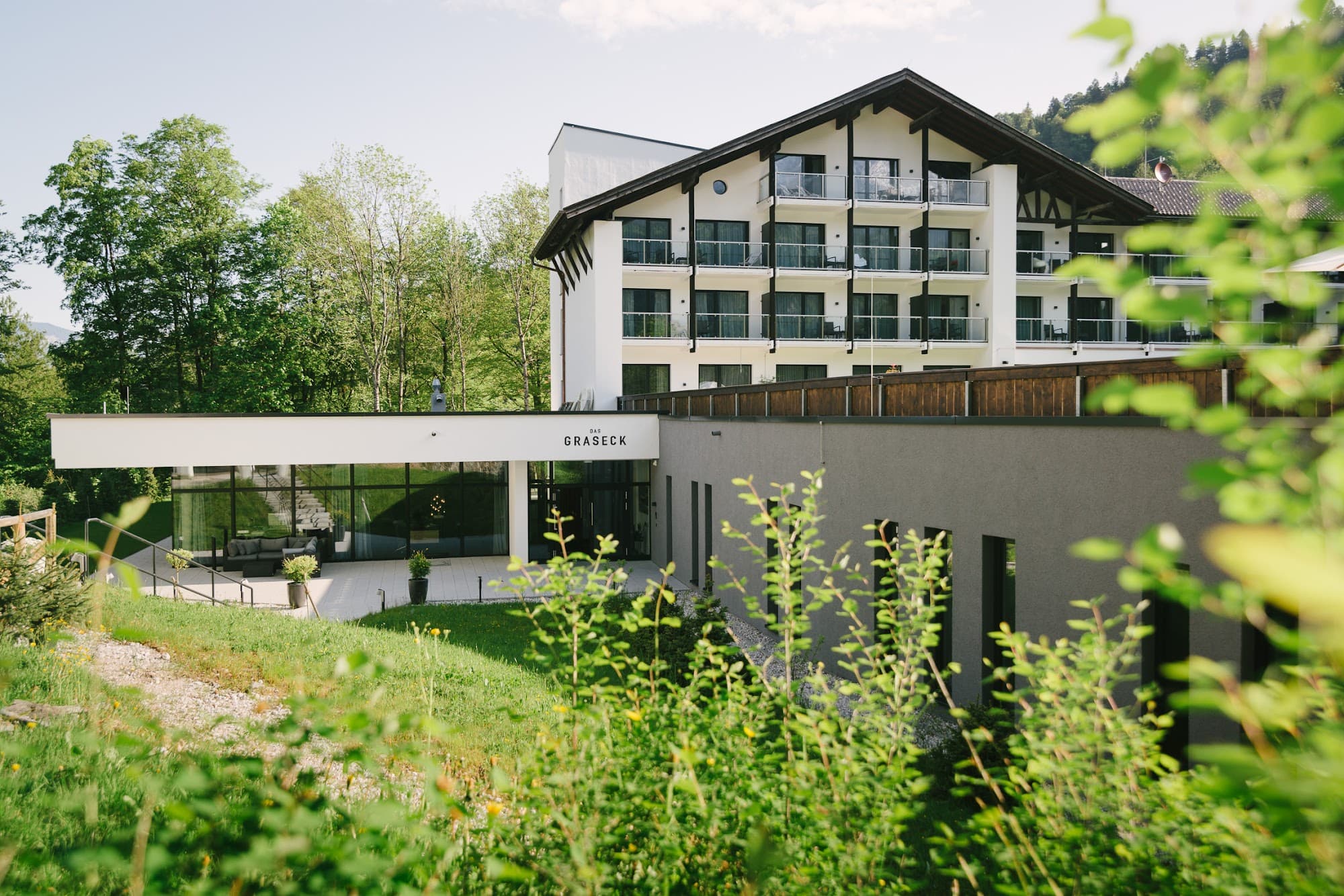 Das Graseck im Sommer: das Hotelgebäude mit Holzbalkonen über dem flachen Spa-Trakt, davor Wiese und Gräser (© Das Graseck, www.das-graseck.de)