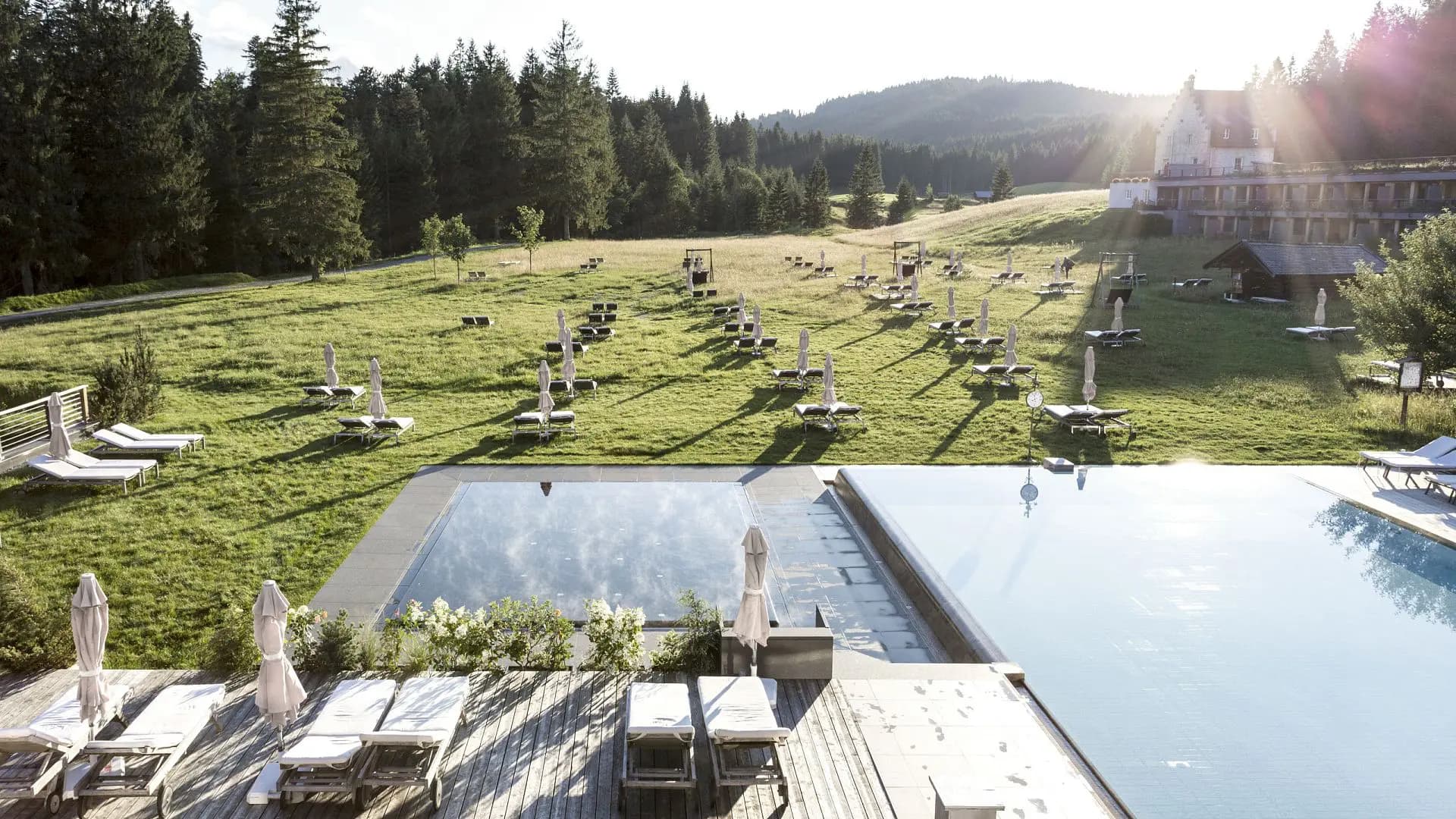 Infinity-Pool und Liegewiese von Das Kranzbach im Sommer, dahinter das englische Landhaus und der Bergwald (© Hotel Kranzbach GmbH / Felix Löchner)