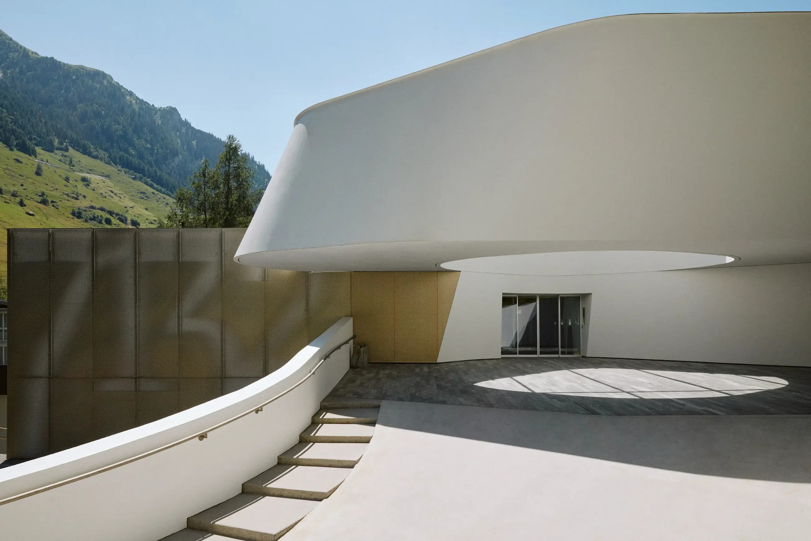 Eingang des 7132 Hotels in Vals — weißer Kurvenbau vor grünem Bergpanorama (© 7132 Hotel / Julien Balmer)