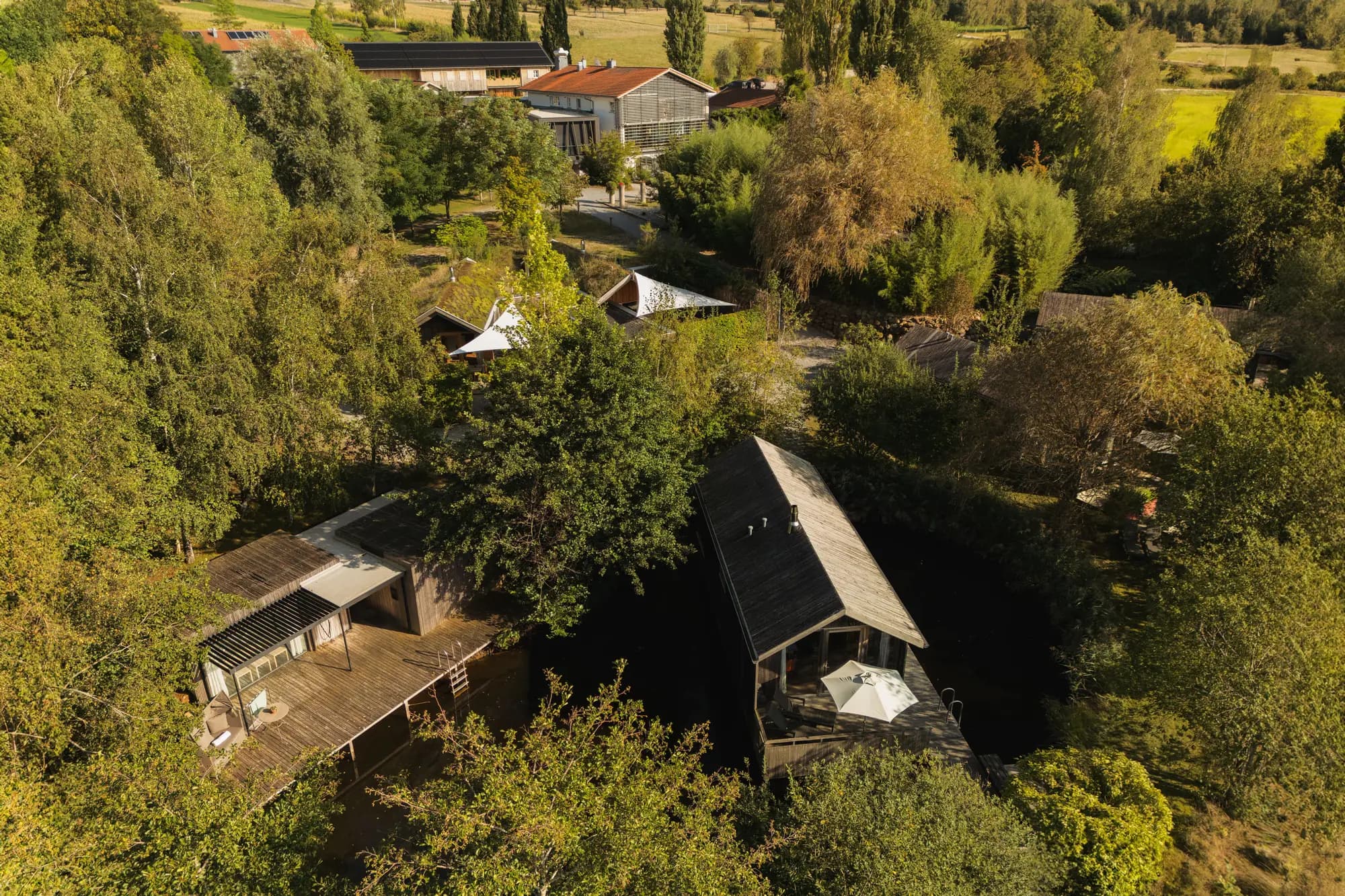 Luftaufnahme des Hofgut Hafnerleiten: zwei dunkle Holzhäuser am Teich zwischen Bäumen, dahinter Sonnensegel, das Haupthaus und Felder (© Hofgut Hafnerleiten / Joschija Bauer)